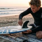 Surfer installing a fin into a surfboard fin box using a fin key on the beach
