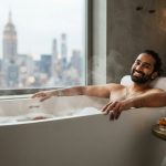 A smiling South Asian man relaxing in a sleek New York City apartment bathtub using an ergonomic bath pillow.